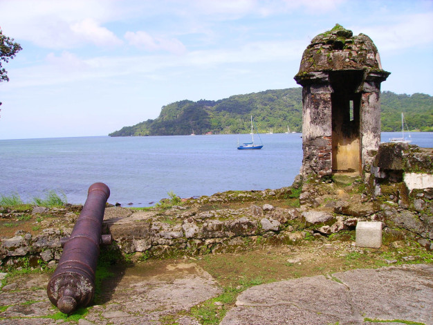 Canon historique donnant sur une baie tropicale avec des voiliers.