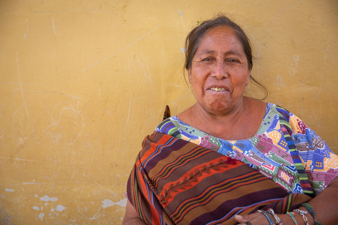 Femme souriante portant un vêtement traditionnel tissé contre un mur jaune.