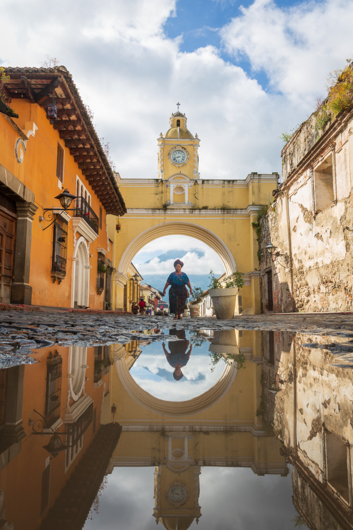 Reflet d'une arche historique dans une flaque d'eau avec une personne qui marche.